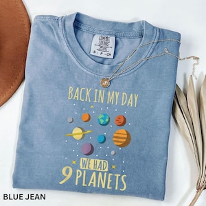 May include: A blue jean Comfort Colors t-shirt with the text "BACK IN MY DAY WE HAD 9 PLANETS" and a graphic of planets. A gold necklace with a pendant is draped across the shirt.