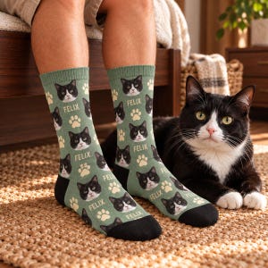 May include: Sage green crew socks featuring a repeating pattern of a black and white cat face, cream-colored paw prints, and the name FELIX printed in white text. The socks have solid black heels and toe sections.
