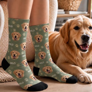Könnte beinhalten: Ein Paar olivgrüne Socken mit einem sich wiederholenden Muster aus Golden Retriever-Gesichtern, weißen Pfotenabdrücken und dem Namen MILO in weißer Schrift. Die Socken haben einfarbig schwarze Fersen und Zehenbereiche.