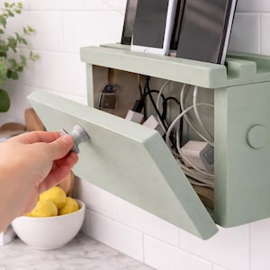 Può includere: Una stazione di ricarica in legno verde chiaro con una porta a cerniera, montata su una parete piastrellata bianca. La porta si sta aprendo per rivelare un groviglio di cavi di ricarica e adattatori di alimentazione. Diverse tavolette sono negli slot superiori.
