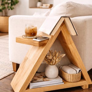 May include: Wooden side table with a triangular design. A glass of iced coffee, a smartphone, and a book rest on the top shelf. The lower shelf holds a white vase with dried flowers, a woven basket, and books.