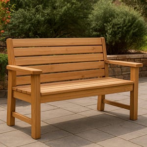 Puede incluir: Un banco de jardín de madera con asiento y respaldo de listones. El banco tiene reposabrazos y está hecho de madera de color marrón claro. Está situado en un patio de piedra con vegetación de fondo.