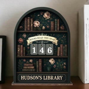 May include: A dark green wooden bookshelf with an arched top, featuring shelves filled with miniature books and floral accents. The text "BOOKS READ THIS YEAR" and "HUDSON'S LIBRARY" are displayed. The number "146" is prominently displayed.
