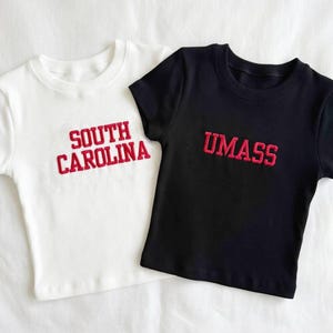 May include: Two short-sleeved t-shirts, one white and one black. The white shirt has "SOUTH CAROLINA" embroidered in red, while the black shirt displays "UMASS" in red. Both shirts are ribbed and have a crew neck.