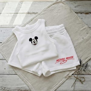 May include: White baby outfit set featuring a tank top with an embroidered Mickey Mouse face and matching shorts. The shorts have "Mickey Mouse" printed in red. The set is displayed on a neutral-colored cloth.