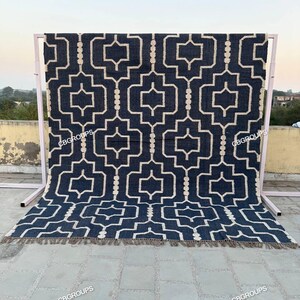 Puede incluir: Una alfombra con estampado geométrico azul marino y crema con borde de flecos. La alfombra se exhibe sobre un marco de metal rosa claro. El patrón geométrico es un diseño repetitivo de formas entrelazadas.