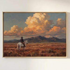 May include: A framed landscape painting depicts a cowboy on a white horse in a vast, arid landscape. The sky is filled with dramatic, orange-hued clouds. Mountains are visible in the distance, and the artwork is set in a gold frame.