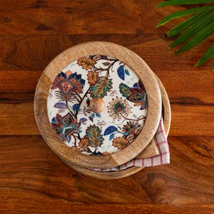 May include: Wooden serving bowl with a floral patterned lid. The lid features a white background with colourful flowers and leaves in shades of blue, orange, and green. A small wooden knob is in the centre. A checkered cloth is visible inside the bowl.