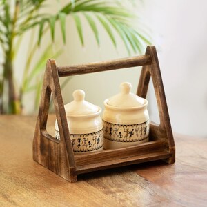 May include: A wooden serving tray with a handle holds two cream-coloured ceramic jars with lids. The jars feature a black and white tribal design around the base. The tray and jars are set on a wooden surface.