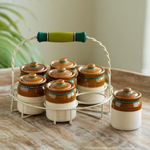 May include: A set of seven ceramic spice jars with lids, in a cream and brown colour scheme, arranged in a white metal carrier with a green and tan handle. The jars have a decorative green band.