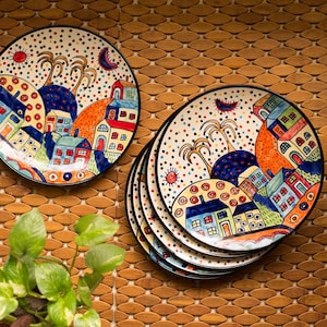 May include: A collection of decorative ceramic plates, each with a vibrant village scene. The plates have a black rim and a whimsical design featuring houses, trees, and suns in blue, orange, red, and yellow. The plates are stacked on a wooden surface.