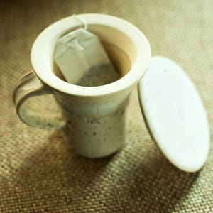 May include: A cream-coloured ceramic mug with a handle, containing a tea bag. The mug has a wide opening and a matching lid resting beside it. The mug has a rustic, handmade appearance, suggesting a cosy, artisanal aesthetic. Ideal for enjoying a warm beverage.
