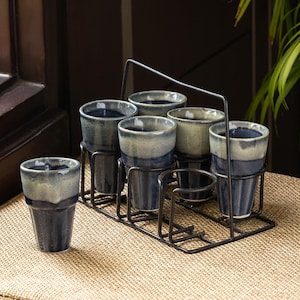 May include: Six ceramic cups with a dark blue and light blue glaze, presented in a black metal rack. One cup is separate. The cups have a textured finish, and the rack includes a handle. The cups are on a tan surface.