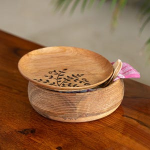 May include: A wooden bowl and lid set with a natural finish. The lid features a black leaf design. A pink and white striped cloth is visible inside the bowl. The set is on a wooden surface.