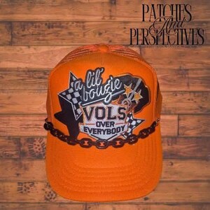 May include: Orange trucker hat with the text "a lil bouzie VOLS over everybody" in white, black, and gold. The hat features a black chain detail and a checkered flag design.