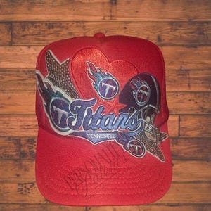 May include: Red baseball cap featuring the Tennessee Titans logo. The logo includes the team name, a football helmet, and a star. The cap has a mesh back and a curved brim.