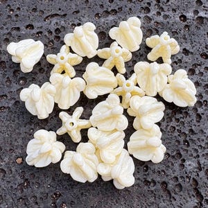 May include: A collection of off-white, flower-shaped beads scattered on a dark, textured surface. The beads vary in design, with some resembling open flowers and others more compact, bud-like shapes. The color is a creamy, neutral tone.