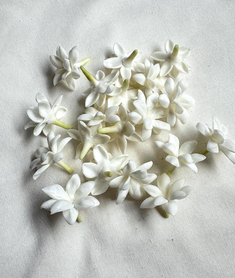 May include: A collection of white jasmine flowers with multiple petals and green stems, scattered on a white fabric surface. The flowers are in various stages of bloom, showcasing their delicate structure.
