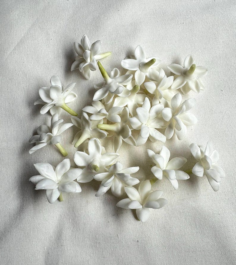 May include: A collection of white jasmine flowers with star-shaped petals and green stems, arranged on a white fabric surface. The flowers are in various stages of bloom, showcasing their delicate structure.