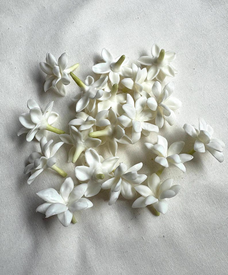 May include: A collection of white jasmine flowers with green stems, arranged on a white fabric surface. The flowers have multiple petals and a star-like shape, creating a delicate and natural aesthetic.