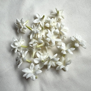 May include: A collection of small, white jasmine flowers with green stems. The flowers are clustered together, showcasing their delicate, multi-petaled structure. The background is a plain, off-white fabric, providing a neutral backdrop for the floral arrangement.