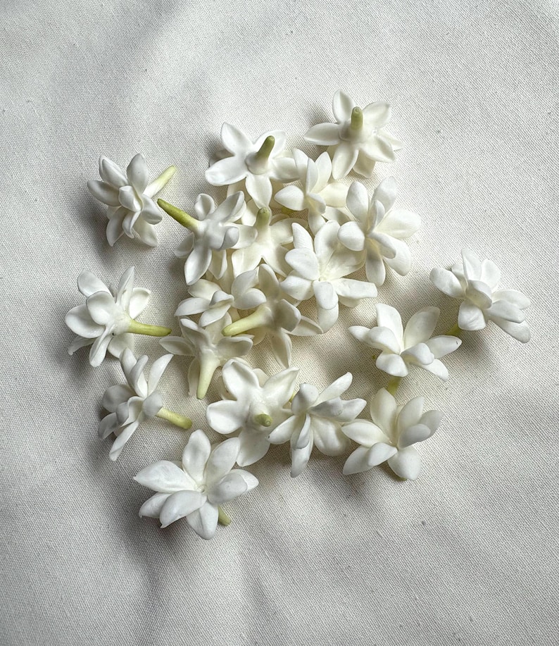 May include: A collection of small, white jasmine flowers with green stems. The flowers are clustered together, showcasing their delicate, multi-petaled structure. The background is a plain, off-white fabric, providing a neutral backdrop for the floral arrangement.