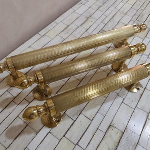 May include: Four brass door pulls with a ribbed center and decorative end caps. The pulls are mounted on round bases. The brass has a warm, golden hue. The hardware is arranged diagonally on a white and beige tiled surface.