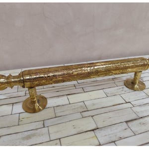May include: A brass door pull with intricate floral and scrollwork designs. The handle is cylindrical with decorative end caps and is mounted on two circular bases. The brass has a polished, golden finish.