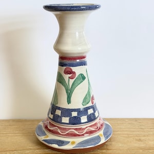 May include: A ceramic candlestick holder with a white body and a flared base. The base features a blue and white checkered pattern, red and blue wavy lines, and floral accents. The top rim is blue, and the holder is decorated with red flowers and green leaves.