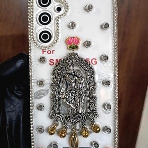 May include: White phone case with silver and gold embellishments. The case features a decorative silver centerpiece with figures, gold charms, and pink lotus flower accents. The text "For SM 5G" is visible.