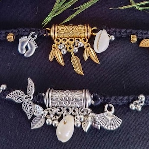 May include: Two black braided cord anklets featuring central metal cylinders. The top anklet has gold-tone leaf charms, a silver-tone baby footprint charm, and a white cowrie shell. The bottom anklet has silver-tone leaf charms, a white shell, and a fan charm.