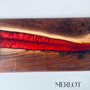 May include: A rectangular serving tray made of walnut wood with a red epoxy resin inlay. The tray has black metal handles on each side and the words "J. Michael Thomas" are engraved on the wood. The word "MERLOT" is printed in black letters below the tray.