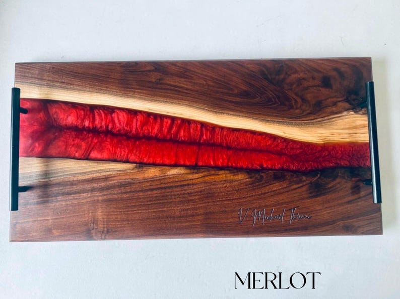 May include: A rectangular serving tray made of walnut wood with a red epoxy resin inlay. The tray has black metal handles on each side and the words "J. Michael Thomas" are engraved on the wood. The word "MERLOT" is printed in black letters below the tray.