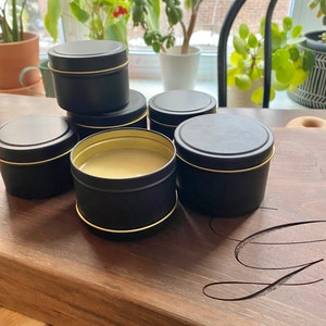 May include: Six small black candle tins with gold rims, arranged on a wooden surface. The tins are filled with a white substance, likely wax.