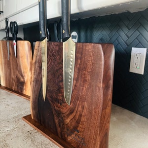 May include: A wooden knife block with a dark brown finish and a natural wood grain pattern. Two gold-handled knives with black blades are mounted on the block. The block has a small wooden base and the text "The Corner Butcher" is engraved on the front.