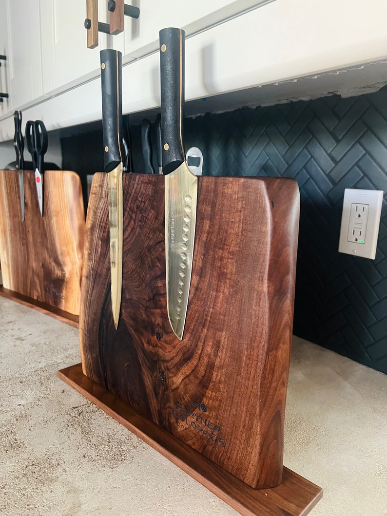 May include: A wooden knife block with a dark brown finish and a natural wood grain pattern. Two gold-handled knives with black blades are mounted on the block. The block has a small wooden base and the text "The Corner Butcher" is engraved on the front.