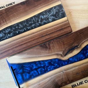 May include: Two wooden serving trays with epoxy resin inlays. The top tray has a black and white epoxy inlay and the text "The Loprieno Family" engraved on the wood. The bottom tray has a blue epoxy inlay and the text "The Hebert Family Est. 2018" engraved on the wood. The top tray is labeled "GUN METAL GREY" and the bottom tray is labeled "BLUE CORAL".