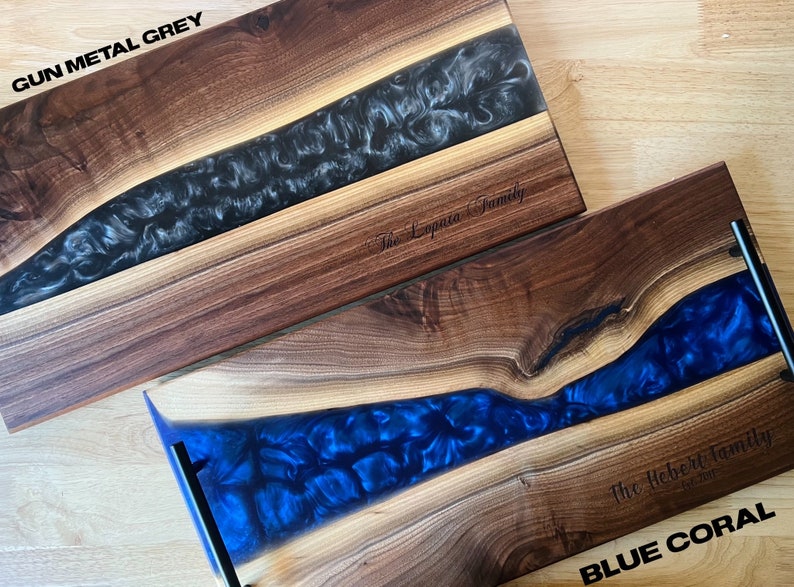 May include: Two wooden serving trays with epoxy resin inlays. The top tray has a black and white epoxy inlay and the text "The Loprieno Family" engraved on the wood. The bottom tray has a blue epoxy inlay and the text "The Hebert Family Est. 2018" engraved on the wood. The top tray is labeled "GUN METAL GREY" and the bottom tray is labeled "BLUE CORAL".