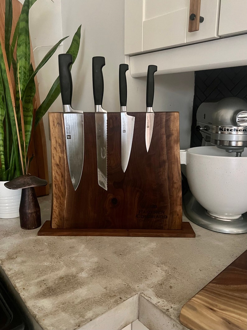 May include: A wooden magnetic knife holder with a dark brown finish. Five silver kitchen knives with black handles are attached to the holder. The holder has the text "Faster Company" engraved on it.