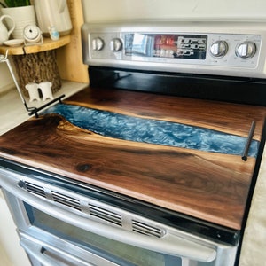 May include: A walnut wood cutting board with a blue epoxy river design. The board is placed on top of a double oven.