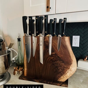 May include: A double-sided knife block made of dark wood with a natural edge. The block is holding a set of eight knives with black handles. The text "DOUBLE-SIDED KNIFE BLOCK" is printed on the counter below the block.