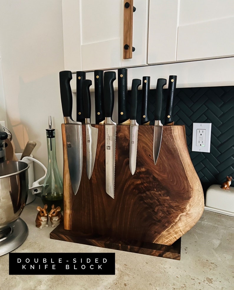 May include: A double-sided knife block made of dark wood with a natural edge. The block is holding a set of eight knives with black handles. The text "DOUBLE-SIDED KNIFE BLOCK" is printed on the counter below the block.