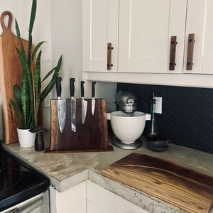May include: A kitchen countertop with a wooden cutting board, a wooden knife holder with five knives, a white stand mixer, and a black and white patterned backsplash.