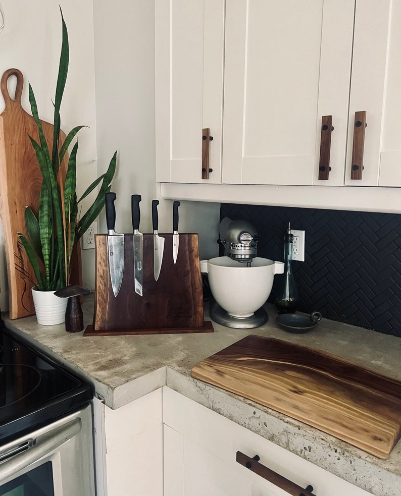 May include: A kitchen countertop with a wooden cutting board, a wooden knife holder with five knives, a white stand mixer, and a black and white patterned backsplash.