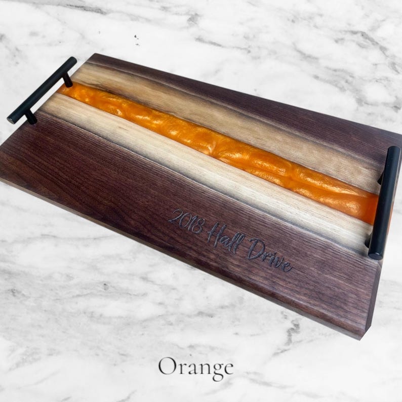 May include: A wooden serving tray with a black metal handle. The tray has a long, thin strip of orange resin running down the center. The tray is engraved with the text "2018 Hall Drive".