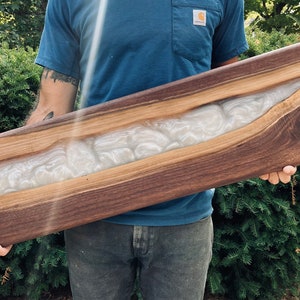 May include: A long, rectangular wooden serving board with a white epoxy river running through the center. The wood has a rich brown color and a natural grain pattern.