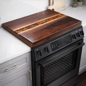 May include: A rectangular wooden stove cover with a dark brown finish and a lighter brown, textured stripe down the center. The cover is placed on top of a black electric stove with control knobs and a digital display.
