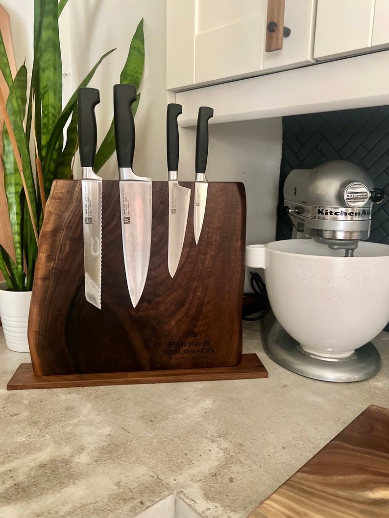 May include: A wooden magnetic knife holder with four silver knives attached. The holder is engraved with the text "Partner Colorado" and "Custom Designs". The holder is sitting on a gray countertop next to a white stand mixer.