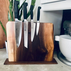 May include: A wooden knife holder with a natural wood grain finish. Five black handled knives are magnetically attached to the holder. The holder has a small wooden base.