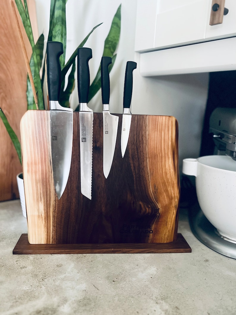 May include: A wooden knife holder with a natural wood grain finish. Five black handled knives are magnetically attached to the holder. The holder has a small wooden base.